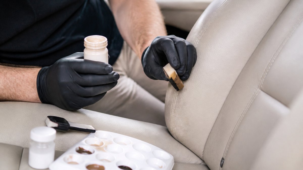 Close-up of a technician's hands applying colour-matched dye to a worn leather car seat bolster, with a small mixing palette and applicator on the seat beside them