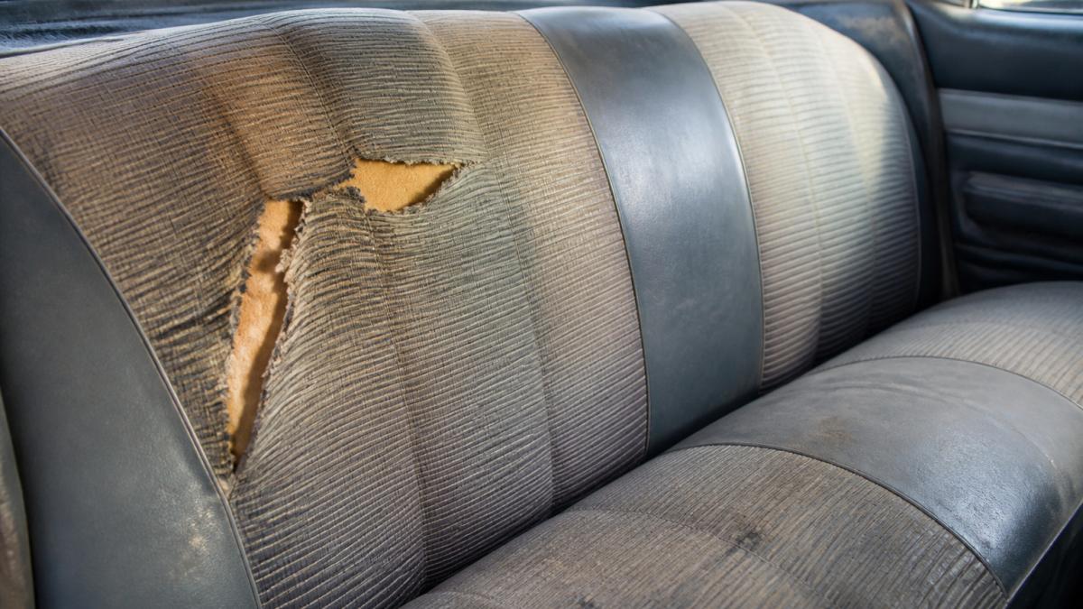 Close-up of classic car interior seat showing various damage types including a visible tear in fabric, worn areas with fading, and discoloration, with natural daylight highlighting damage details